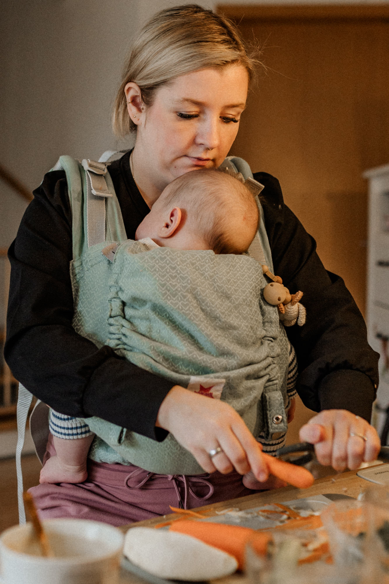 Mama, die mit ihrem Baby in der Trage Karotten schält